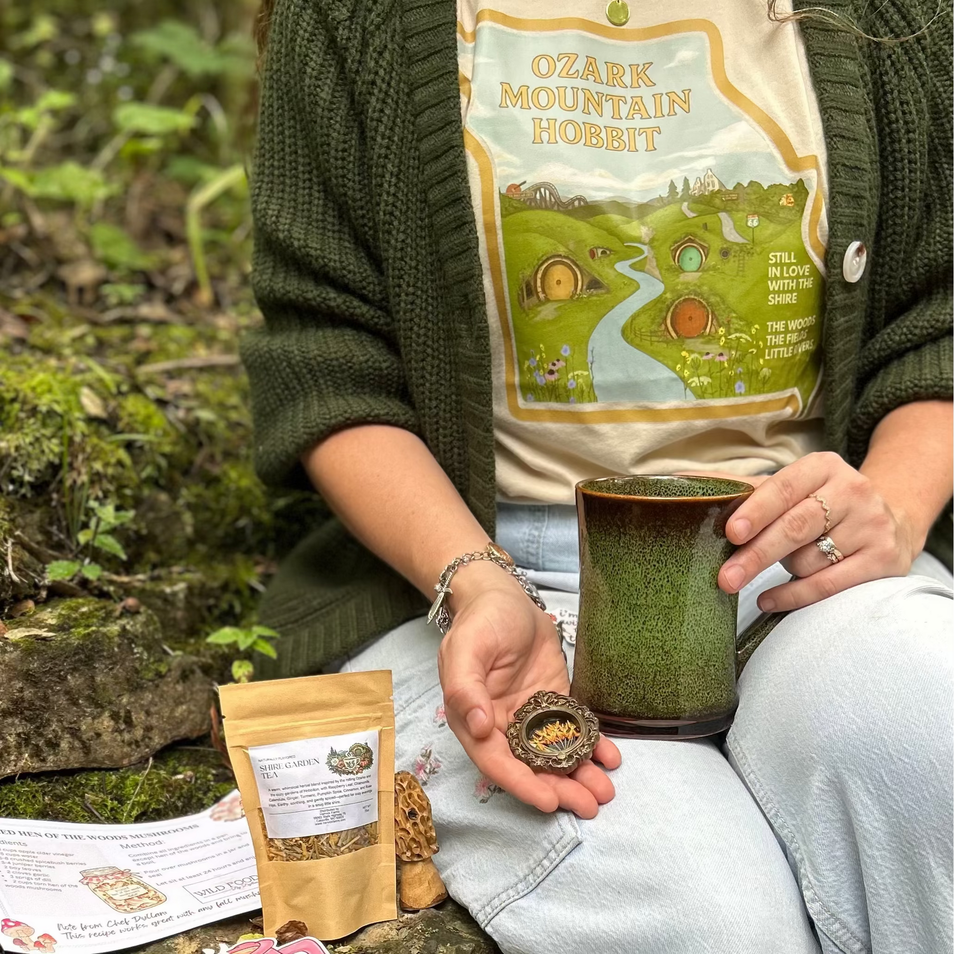 A person sitting outdoors on a mossy stone ledge wearing a cream graphic T-shirt that reads 'Ozark Mountain Hobbit' with a green cardigan and light blue jeans, holding a ceramic mug. A small kraft pouch, a printed card, and a round item are placed on the stone beside them in a forest setting.